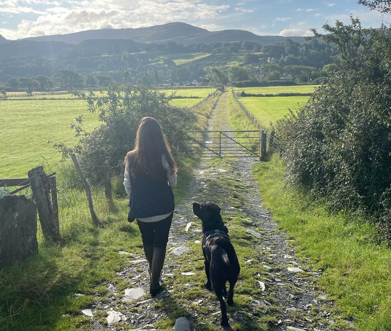 Person walking with a black labrador along a path on a sunny day with hills in the background